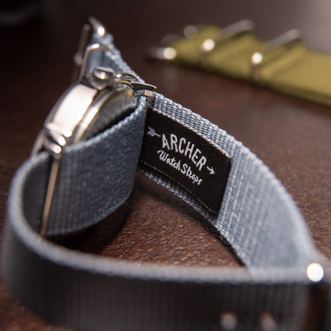 Close-up of a branded woven label tag on a gray military style watch strap through a hardware ring